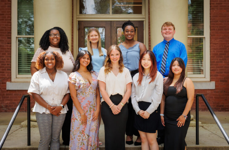 Nine people pose for a group photo on steps outside a brick building with white columns and glass doors.