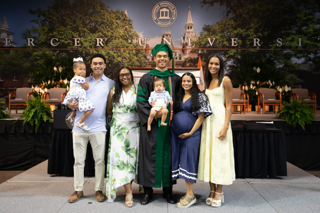 A group of six people stands together indoors at a graduation ceremony, with one person in a green cap and gown.