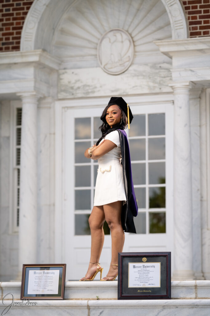 Addison Robinson, in graduation attire, stands on steps in front of building, with two framed diplomas displayed on the ground.