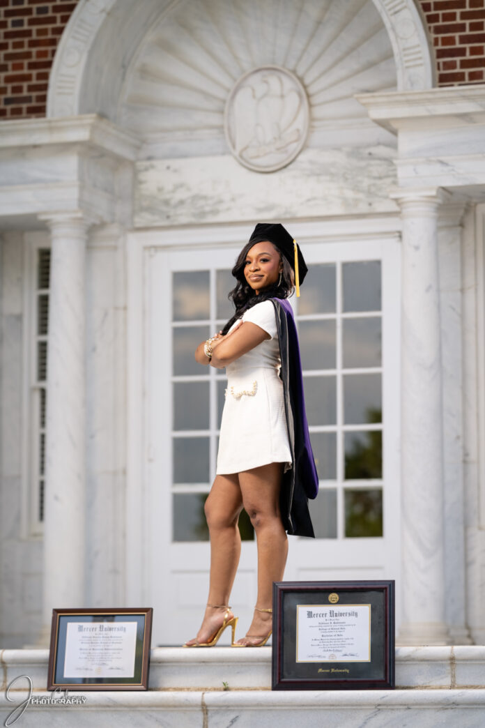 Addison Robinson, in graduation attire, stands on steps in front of building, with two framed diplomas displayed on the ground.