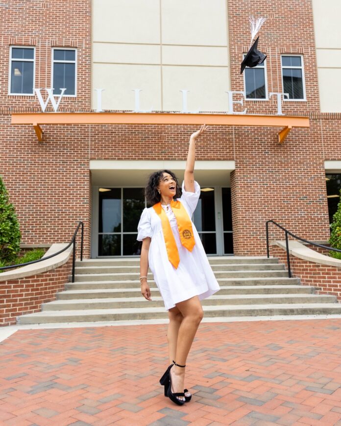 Ashley Boone, in a white dress and orange sash, throws her cap in the air outside the Willet Science Center.