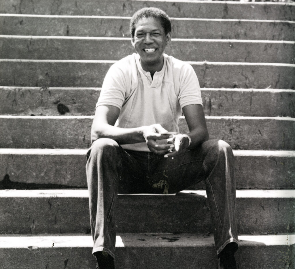 A man sits on outdoor concrete steps, smiling, wearing a light polo shirt and pants, with hands clasped in front.