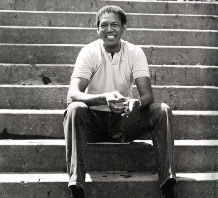A man sits on outdoor concrete steps, smiling, wearing a light polo shirt and pants, with hands clasped in front.