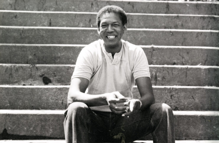 A man sits on outdoor concrete steps, smiling, wearing a light polo shirt and pants, with hands clasped in front.