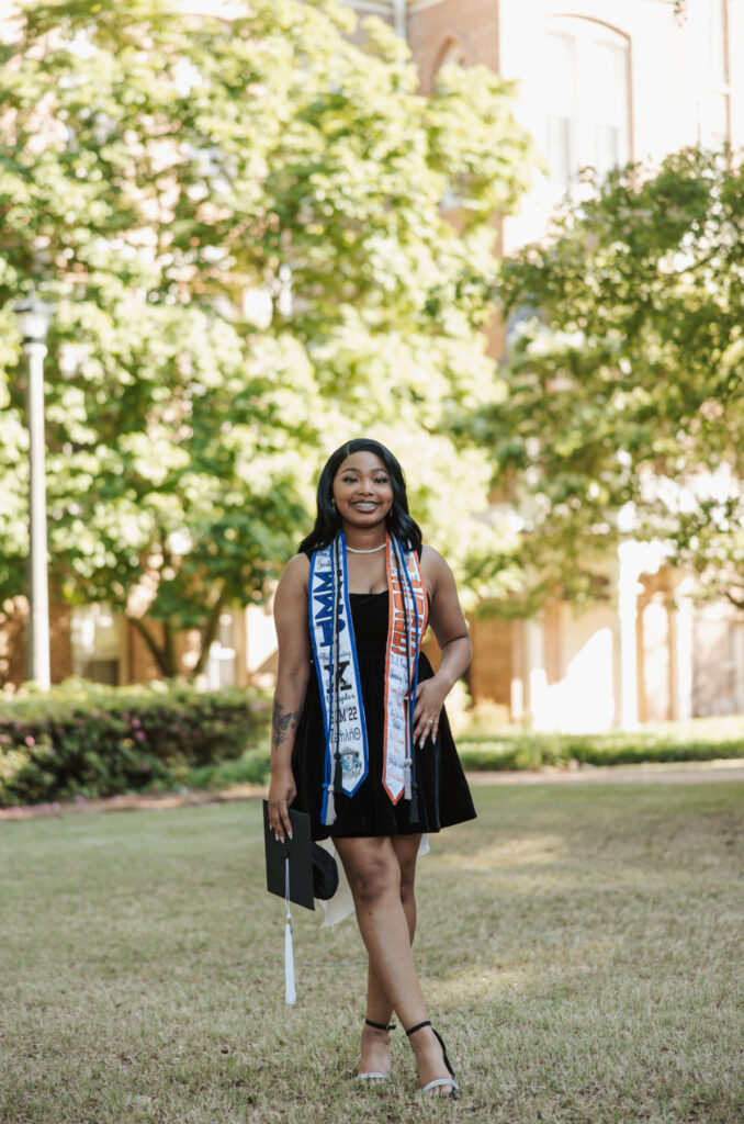 Woman in a black dress stands on grass, smiling and holding a graduation cap, with stoles draped around her neck.