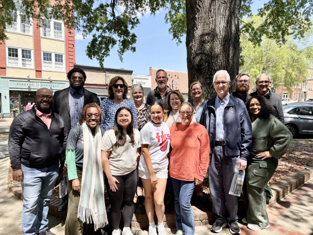 A group of sixteen people poses together outdoors in front of a large tree on a sunny day.