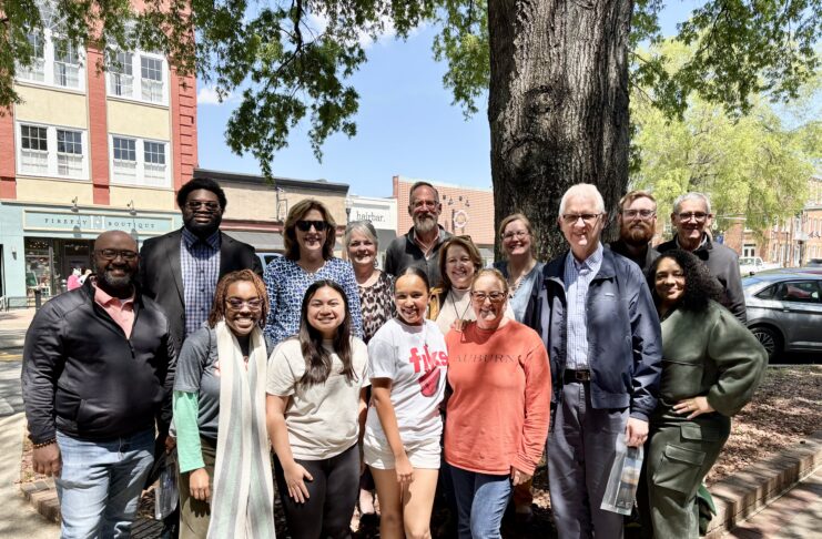 A group of sixteen people poses together outdoors in front of a large tree on a sunny day.