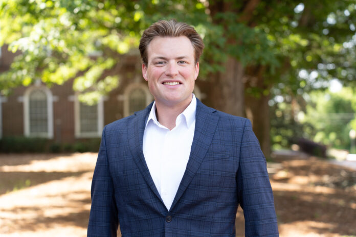 Colby Belland stands outdoors, smiling, with trees and a brick building in the background.