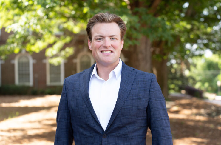 Colby Belland stands outdoors, smiling, with trees and a brick building in the background.