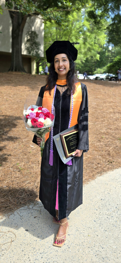 Deepthy Varghese, in cap and gown, holds a bouquet of flowers and a plaque, standing outdoors on a sunny day.