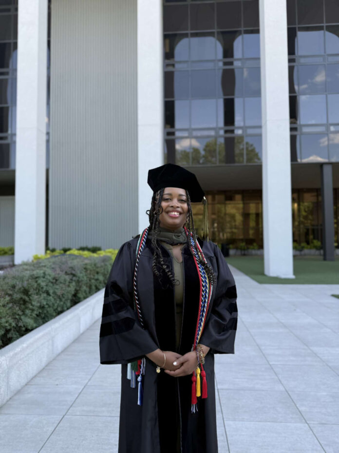 Glennessa Hodge, in a cap and gown, stands smiling outside a modern building.