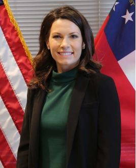 Woman with long brown hair in a dark blazer stands in front of American and state flags, smiling at the camera.