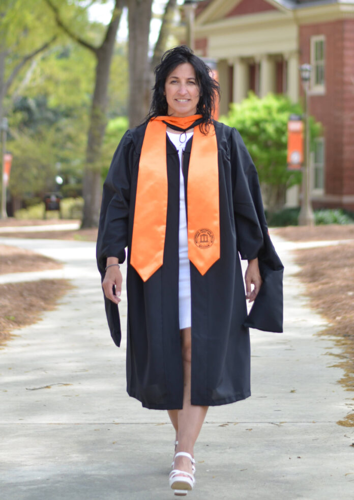 Jennifer Jackson, in a graduation gown and orange stole, walks outside on a sidewalk, smiling at the camera.