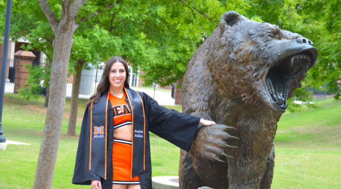 Graduate Spotlight: Kinleigh King Kinleigh King, in a graduation gown and cheerleading uniform, stands by a roaring bear statue.