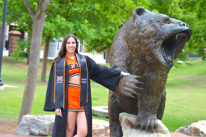 Kinleigh King, in a graduation gown and cheerleading uniform, stands by a roaring bear statue.