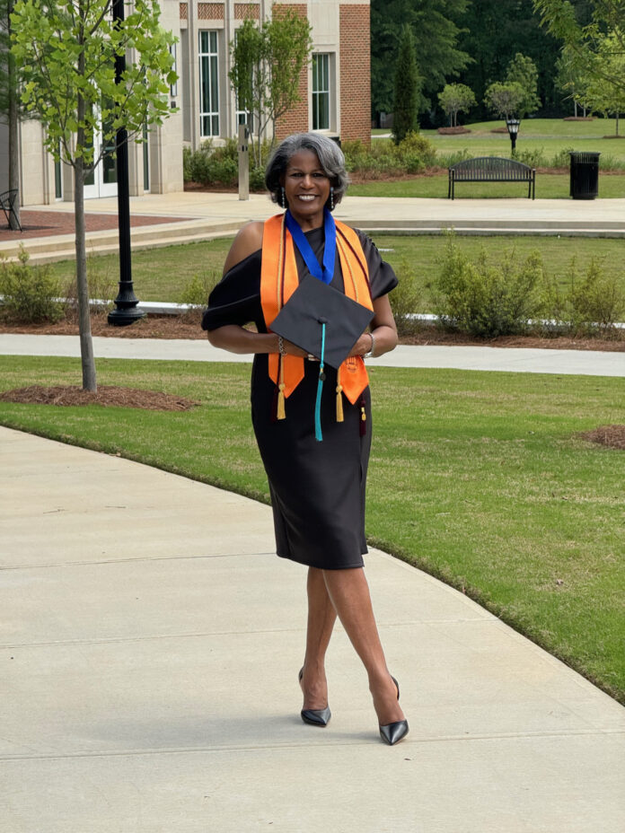 Leidra Dunson, in a graduation outfit, holds a cap and smiles while standing on a sidewalk near a building.