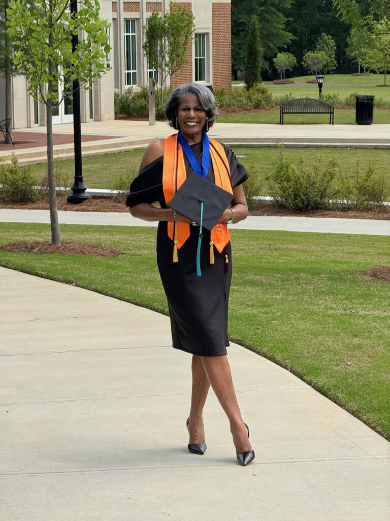 Leidra Dunson, in a graduation outfit, holds a cap and smiles while standing on a sidewalk near a building.
