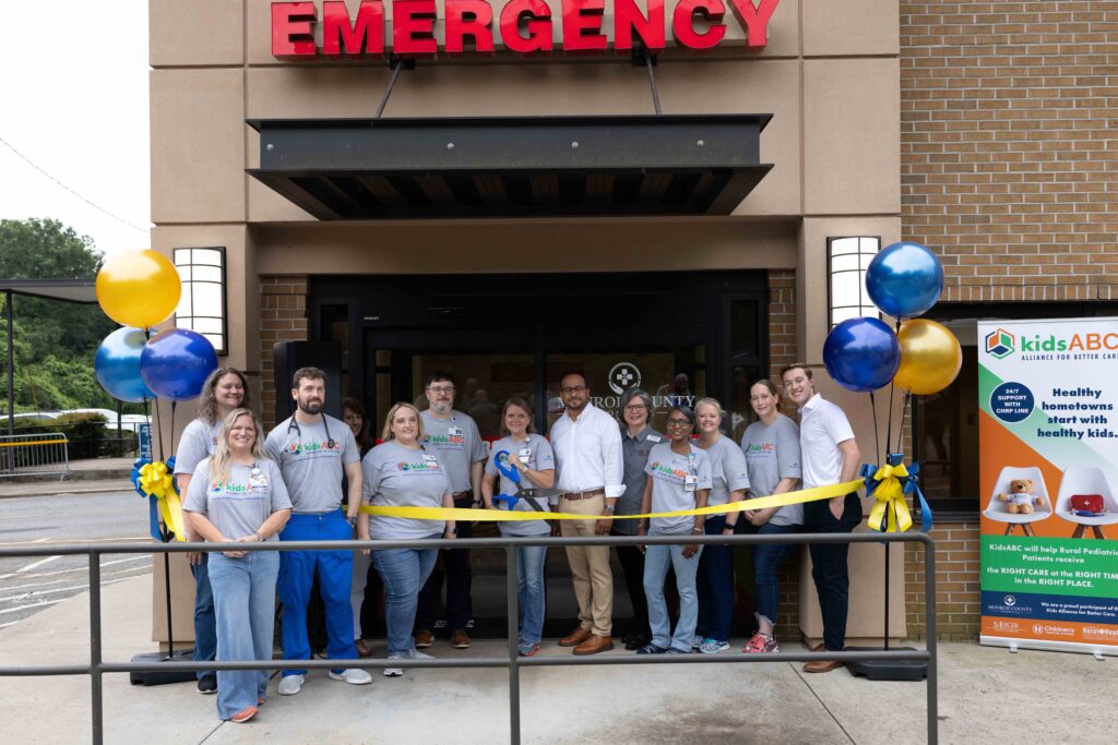 A group of people stands in front of an emergency entrance for a ribbon-cutting ceremony with balloons and a banner.