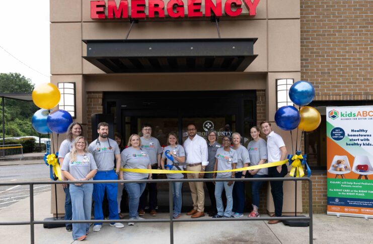 A group of people stands in front of an emergency entrance for a ribbon-cutting ceremony with balloons and a banner.