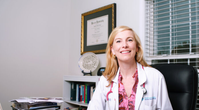 As a pediatrician, alumna loves watching children grow A woman in a white coat sits at a desk in an office with medical papers, books, and a diploma on the wall.