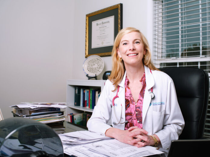 A woman in a white coat sits at a desk in an office with medical papers, books, and a diploma on the wall.