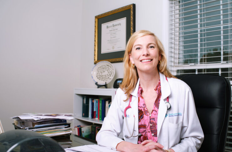 A woman in a white coat sits at a desk in an office with medical papers, books, and a diploma on the wall.