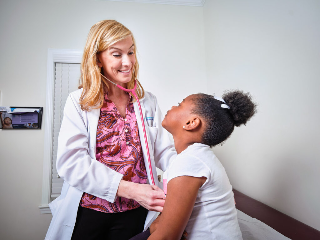 A female doctor uses a stethoscope to listen to a young girl’s chest during a medical exam in a clinic room.