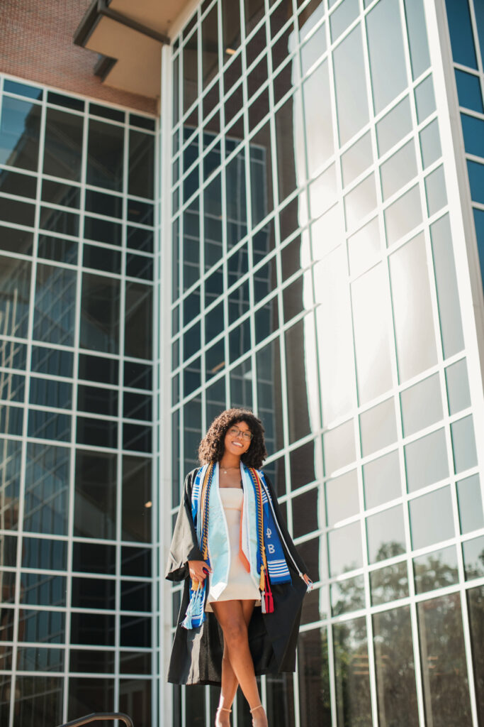 Maya Henry, in graduation attire, stands outside a modern glass building, smiling and posing for a photo.