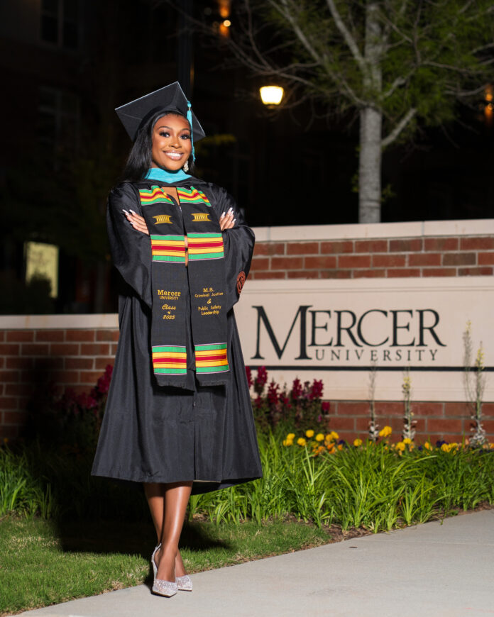 Morgan Taylor, in cap and gown, stands with arms crossed in front of a Mercer University sign at night.