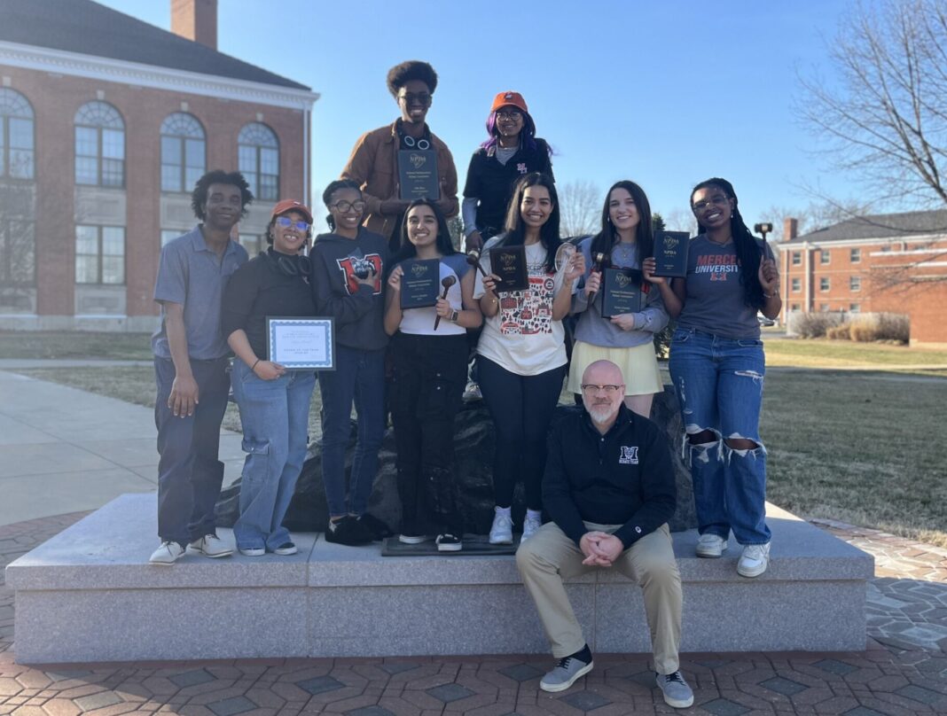 A group of ten people pose on a stone platform outdoors, holding plaques and certificates, with buildings in the background.