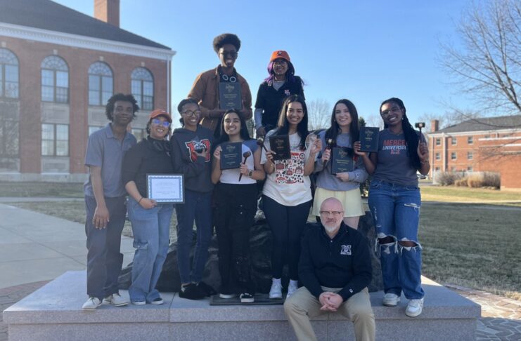 A group of ten people pose on a stone platform outdoors, holding plaques and certificates, with buildings in the background.