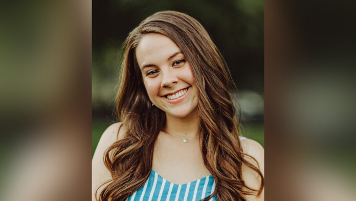 A young woman with long brown hair smiles outdoors, wearing a blue and white striped top.