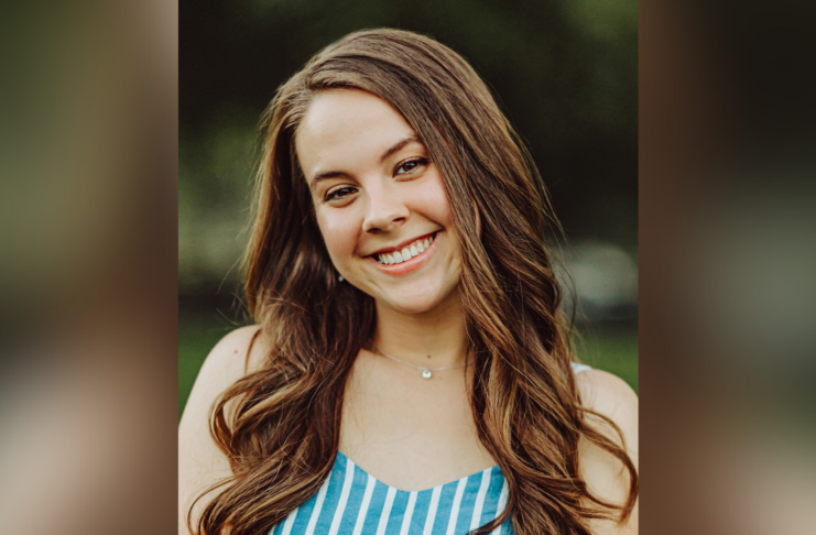 A young woman with long brown hair smiles outdoors, wearing a blue and white striped top.
