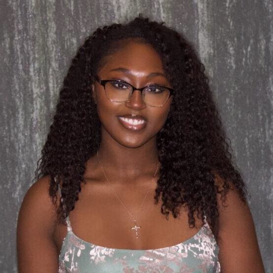 Woman with long curly hair and glasses smiles at the camera, wearing a floral dress and a cross necklace.