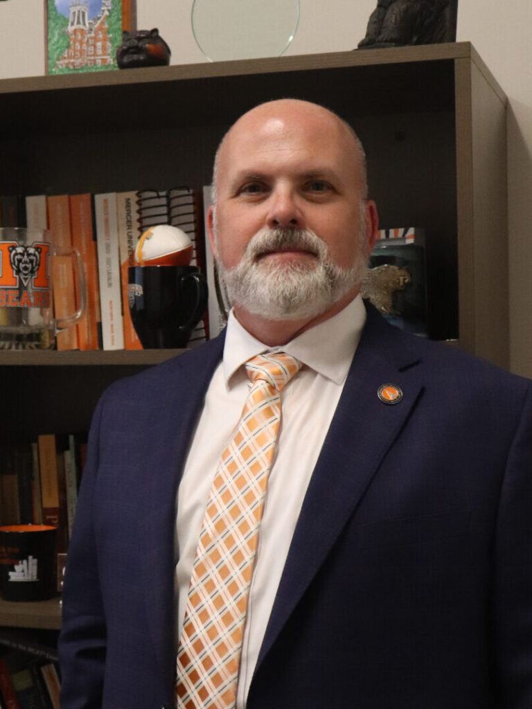 Garland Crawford, wearing a navy blue suit and orange checkered tie, stands in front of a book shelf.