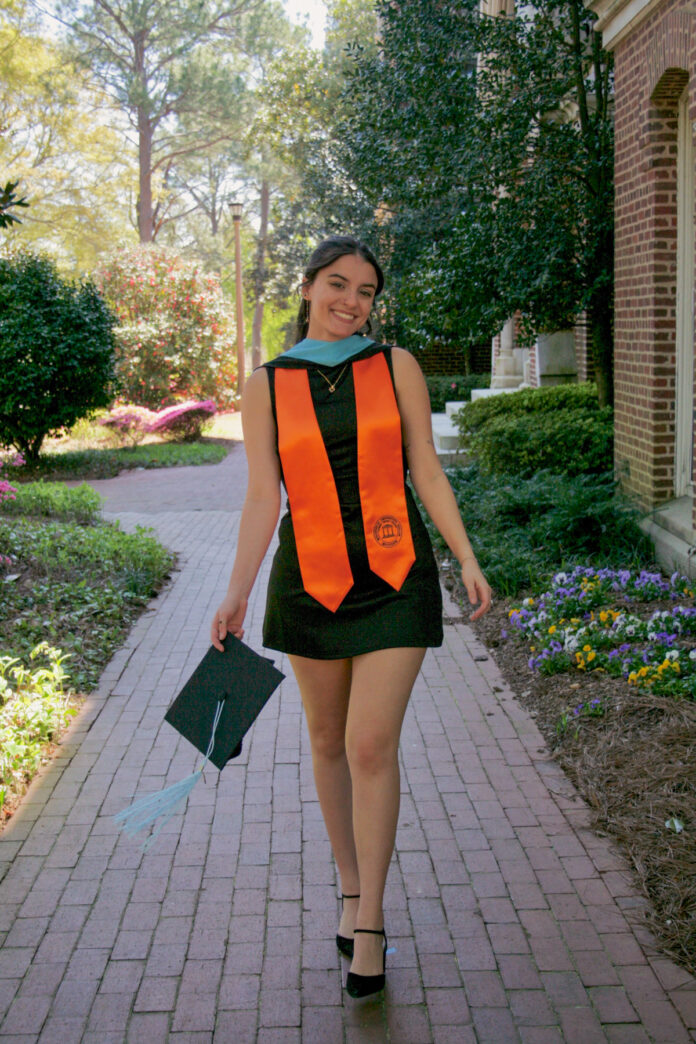 Morgan Goeckel, in graduation attire, walks down a brick path outdoors, holding her cap and smiling.