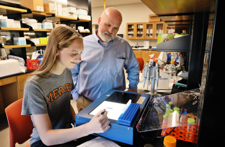 A student and a professor observe lab results on a screen in a science laboratory surrounded by equipment and supplies.