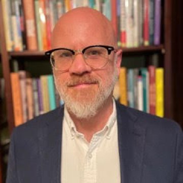 A man with glasses and a beard stands in front of a bookshelf filled with books, wearing a white shirt and a dark jacket.