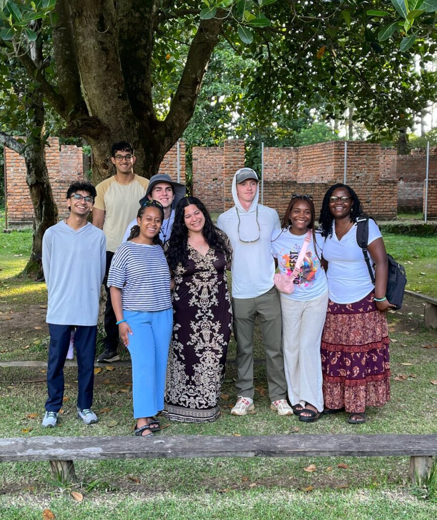 A group of eight people pose together outdoors in front of a large tree and brick wall.