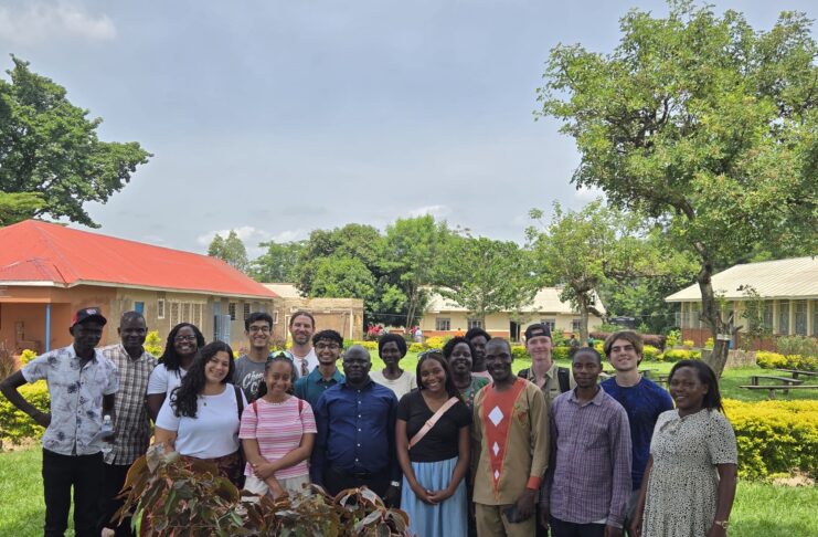 A group of people stands together outdoors on grass, smiling, with buildings and trees in the background.