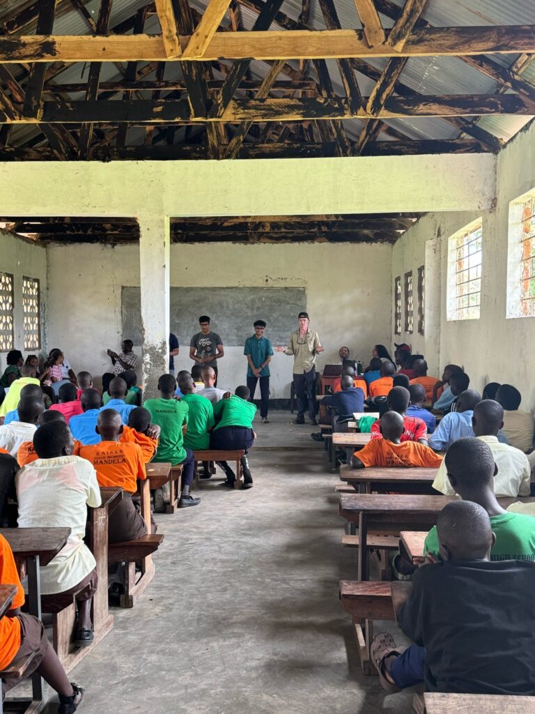Students in colorful uniforms sit in a classroom with three adults standing at the front addressing them.