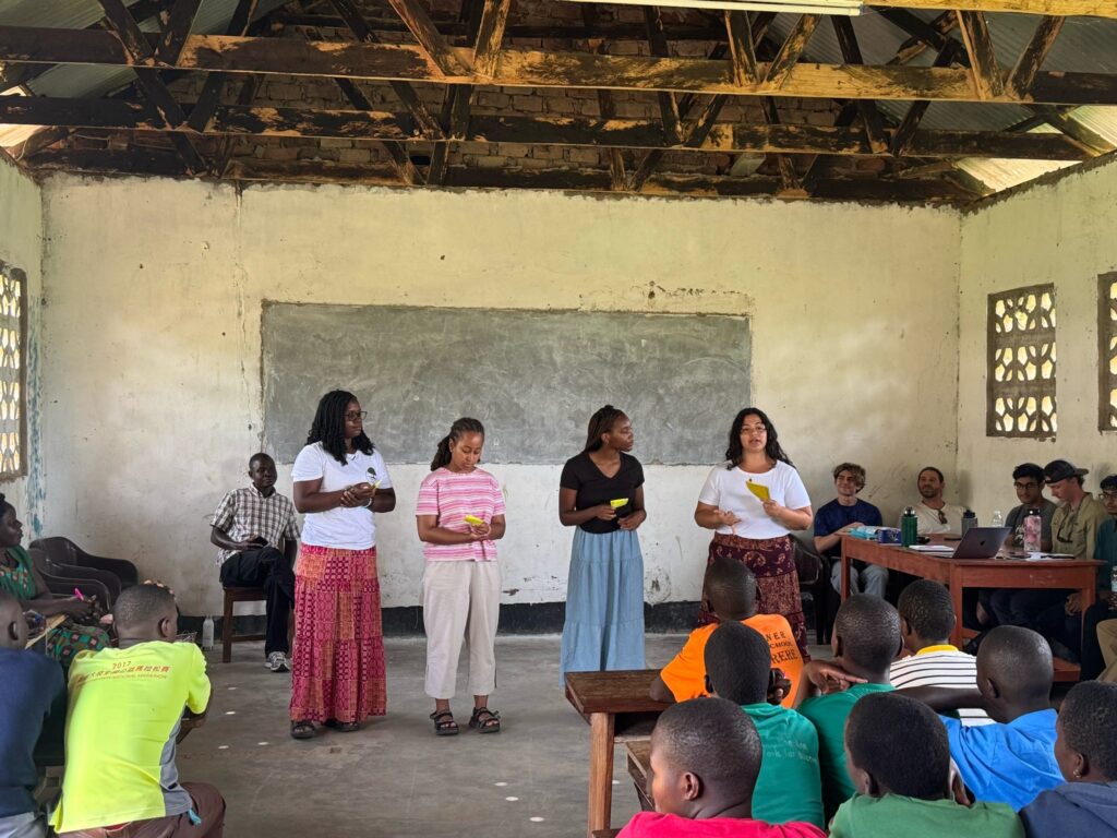 Four women stand at the front of a classroom, speaking to a group of seated children and adults.