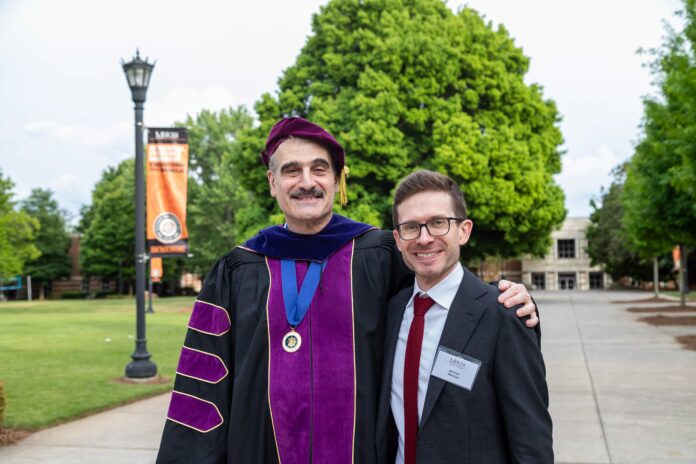 2025 Businessrection-Jamietucker-SM Two men, one in academic insignia and one in costume, stand together outside on a campus with green trees in the background.