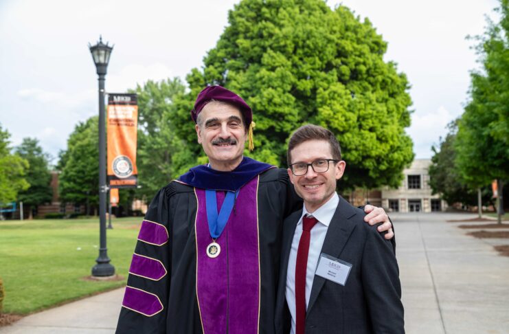 Two men, one in academic regalia and one in a suit, stand together outside on a campus with green trees in the background.