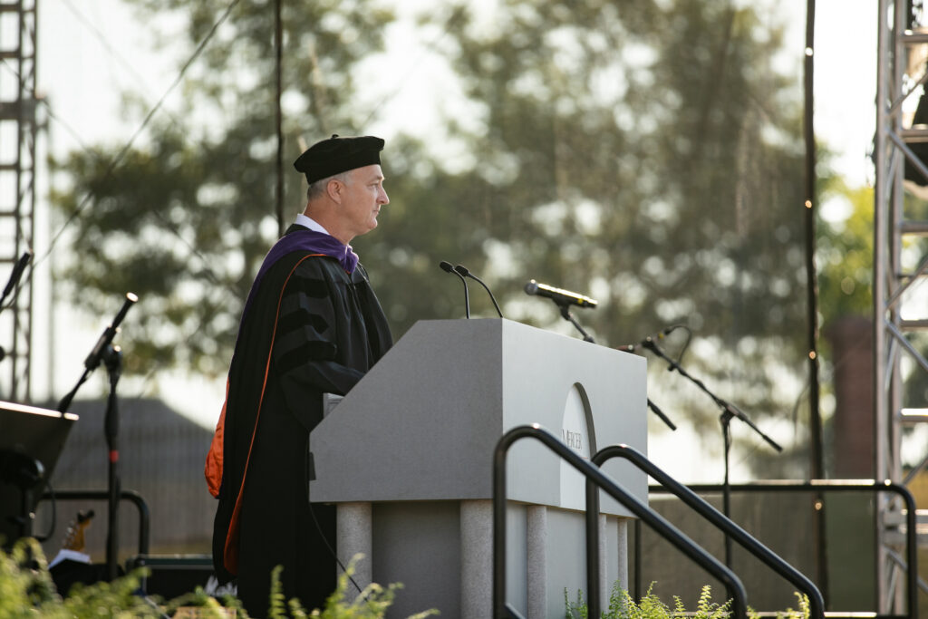 A person in academic regalia speaks at an outdoor podium during a graduation ceremony.