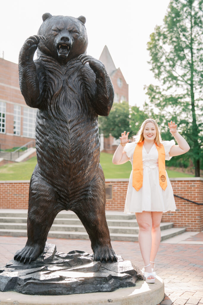 Abby Milam, in a white dress and graduation stole, poses imitating a bear next to a large bear statue outdoors.