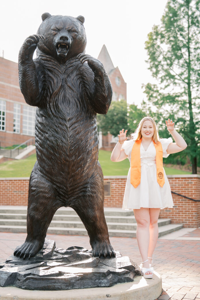 Abby Milam, in a white dress and graduation stole, poses imitating a bear next to a large bear statue outdoors.