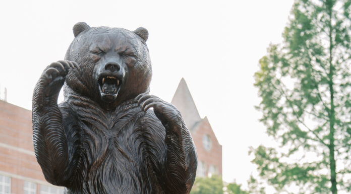 Graduate Spotlight: Abby Milam Abby Milam, in a white dress and graduation stole, poses imitating a bear next to a large bear statue outdoors.