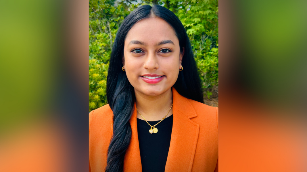 A woman with long dark hair in an orange blazer stands outdoors, smiling slightly, with greenery in the background.