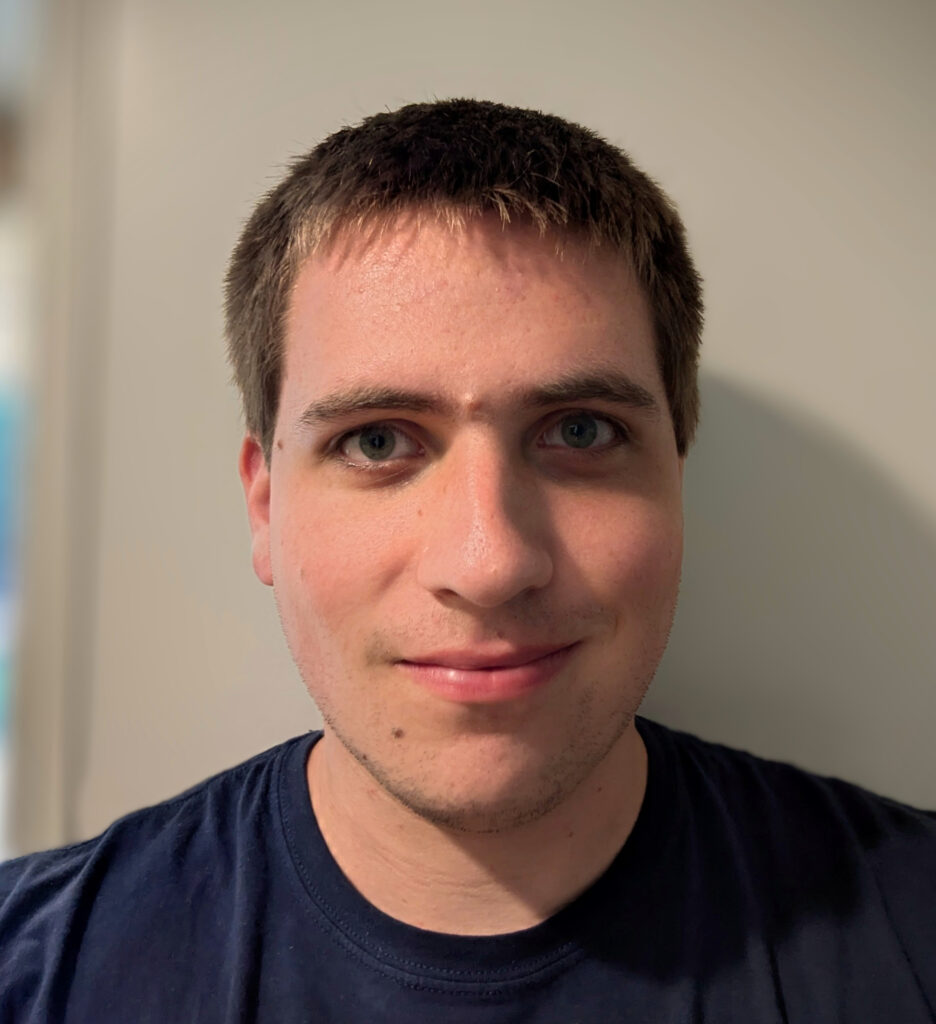 A young man with short brown hair and blue eyes looks at the camera, smiling slightly, against a plain background.